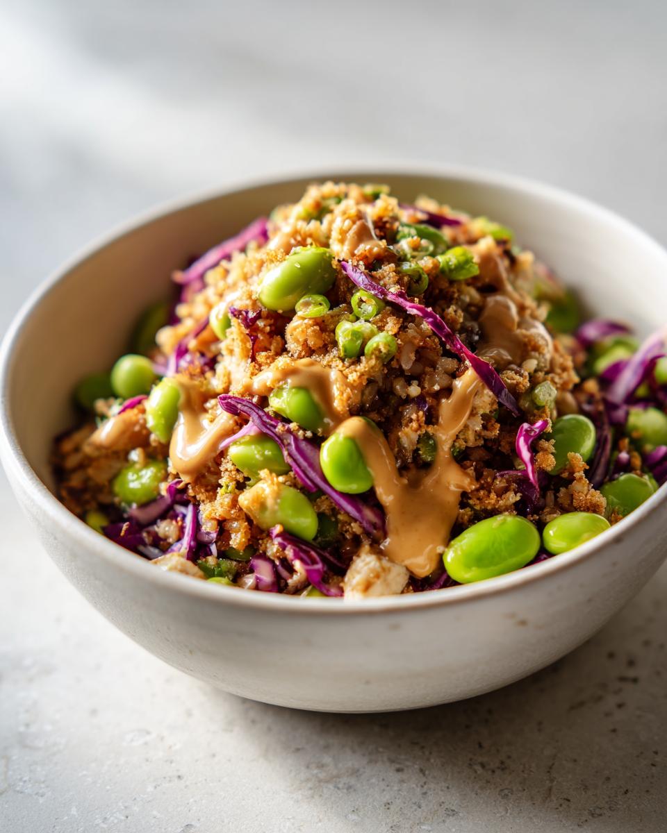 Close-up of a bowl featuring crispy rice salad topped with bright green edamame, shredded purple cabbage, and drizzled peanut sauce.