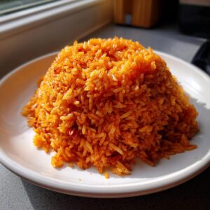 A perfectly mounded serving of vibrant, fluffy jollof rice on a white plate, highlighted by natural sunlight.