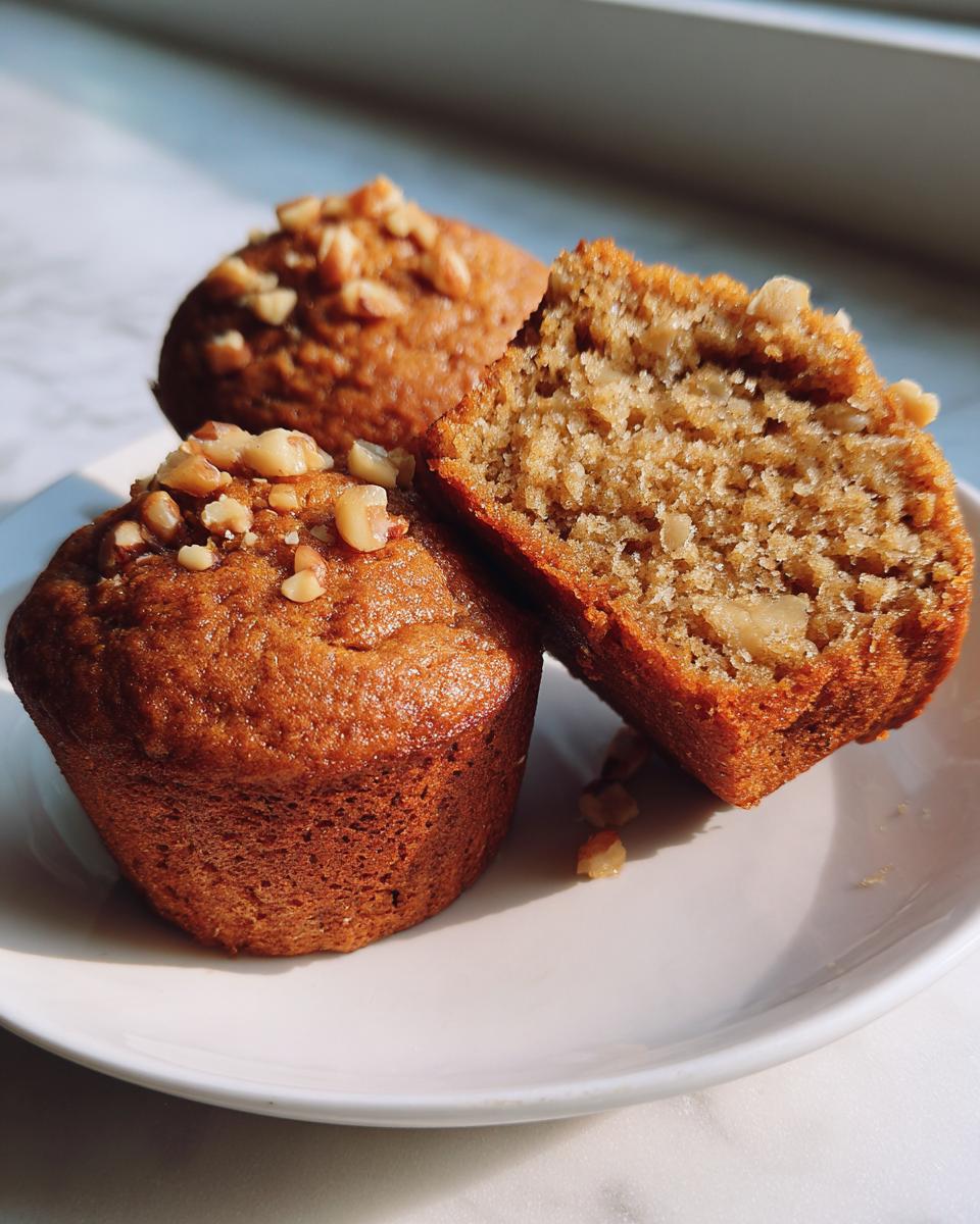 Close-up of two golden brown banana nut muffins on a white plate, one split open showing the moist interior and walnuts.
