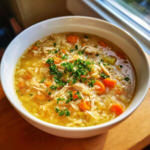 A close-up of a white bowl filled with rich, golden penicillin soup featuring shredded chicken, rice, carrots, and fresh parsley.