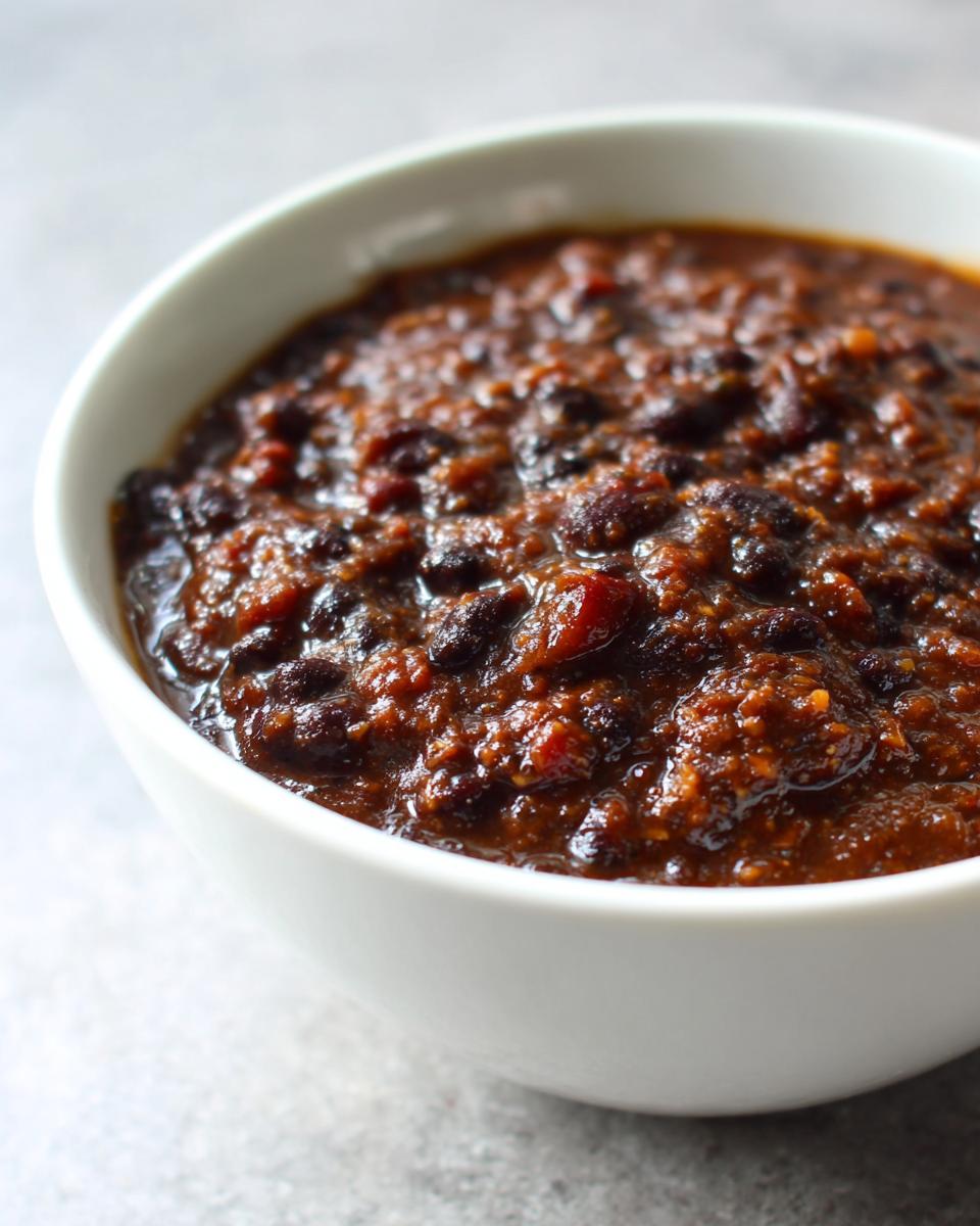 Close-up of thick, rich black bean chili served in a white bowl against a light gray background.