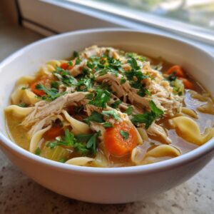 A close-up of a white bowl filled with rich slow cooker chicken noodle soup, topped with shredded chicken, carrots, and fresh parsley.